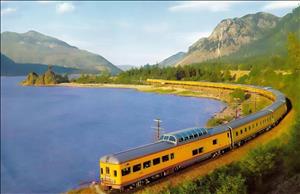 A long yellow passenger train along a large river. The train curves around a bend in a river. On the other side of the tracks are green hillsides. The last car on the train has a glass dome on top. 