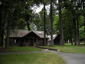 A log cabin in a stand of conifers with paved trails cutting through mowed grass in front of the cabin. 