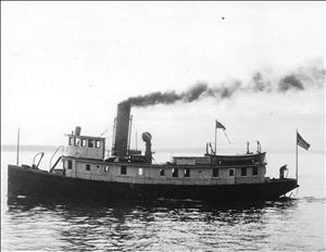 Black and white photo of a small steamboat with dark hull and lighter cabin area 