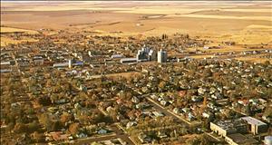 Bird's eye view of a town on a flat prairie. Several blocks of houses and trees lead up to a few city buildings and factories. 