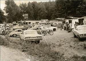 People stand in front of small shacks in a parking lot full of cars and motorcycles