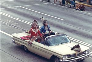 Two indigenous men in traditional garb and head pieces ride on the back of a convertible Cadillac