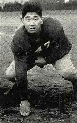 A young Asian man in a football quarterback pose with one fist on the ground. He is standing on a sports field. 