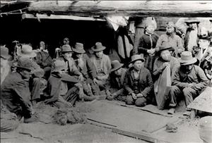 A large group of indigenous people in hats and suit coats kneel and sit in a semi circle next to a wooden structure. 