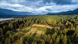 Aerial view of large hotel building in clearing on slope otherwise covered with large evergreen trees, rising to mountains in distance, and dropping to a small stretch of river at left 