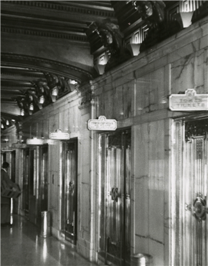 A shiny interior marble wall with four ornate elevator doors each with a sign above it. 