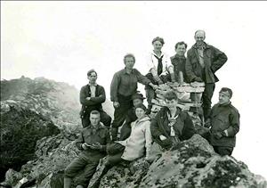 Men and women in early 20th century hiking clothes sit and stand on a steep rocky peak