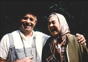 An elderly man with his arm around the shoulder of an elderly woman. Both are laughing. The woman has a scarf on her head and the man is wearing striped overalls. There is green forest behind them. 