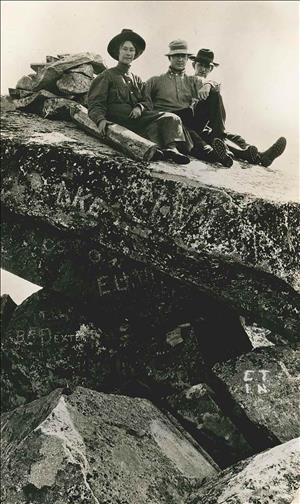 A woman and two men seated on top of a pile of large rocks in early 20th century hiking clothes