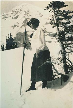 A woman in thick skirts, sweaters, boots, and a hat stands in the snow with a walking stick. A snowy peak in the background.  