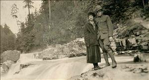 A white man in a military uniform stands with his arm around a white woman in a winter coat and hat. They are standing on a rock beside a rushing river in the woods