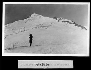 A woman in winter clothing stands just below the summit of a snow-covered mountain peak