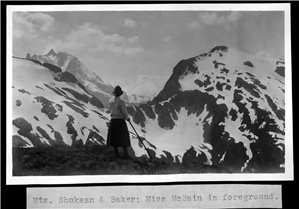 A woman with a hiking stick has her back to the camera and is looking out at a snow-covered mountain peak