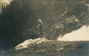 A woman in early 20th century hiking boots stands on a rock in the clearing in the forest. There is snow on the ground. She is facing the forest and looking back over her shoulder at the camera with her hair blowing in the wind. 