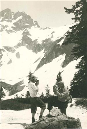 A man and woman face each other with cameras held up to their faces. They are standing on a snowy bank and a snow capped mountain peak looms in the background. 