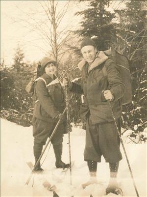 A man and a woman stand in the snow smiling. They are wearing winter clothing, large backpacks, knee high boots, and snowshoes. They are holding hiking poles. 