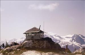 A one room wooden structure with windows on all sides perched on a rock. In the distance are snow covered peaks. 