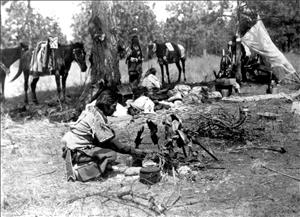 An indigenous person sits on the ground stoking a fire. Pieces of fish are perched over the fire on sticks. In the background are other indigenous people and some horses. 