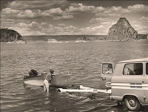 A person stands in the water next to a small boat that sits on a trailer that is attached to a van that has backed up to the water's edge. Another person stands where the trailer connects to the van. Across the body of water are large rocky bluffs and dramatic clouds. 