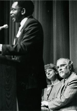 A black man stands at a podium while a white man and white woman watch from their seats behind him. 