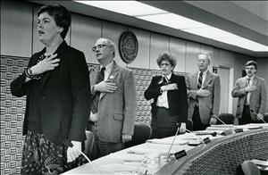 Five people in a row each holding their right hand over their heart and facing the same direction. They are wearing business attire and standing at a long, curved podium. 