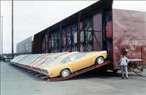 Yellow, green, and red cars, all of the same model, are loaded onto a ramp attached to a rail car. Inside the rail car are more of the same car stacked vertically inside the car. A person in a light blue suit inspects the front of the train car. 