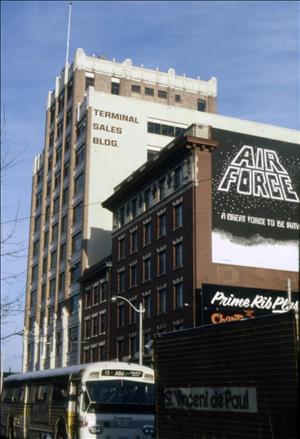 A brown brick building several stories high with the words terminal sales building painted on the side. Two smaller red brick buildings are in the foreground. A billboard says air force a great force to be with. A yellow and white public bus passes. the route sign on the front of the bus says fifteen alki harbor island. 