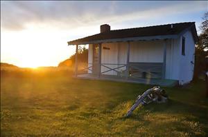 A small, white, wooden cabin on a grassy bluff with a sunset behind it