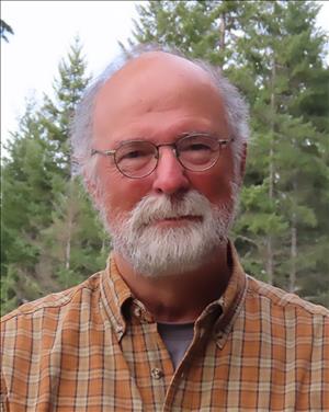 An older white man with round eye glasses and a white beard in an orange and brown plaid shirt. There are evergreen trees behind him. 