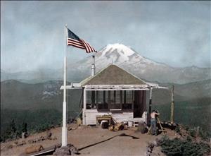 A one room structure with windows on all sides looks out on a large snow capped peak. There is an American flag on a flagpole beside the cabin. A person is standing outside looking through equipment on a wooden tripod. 