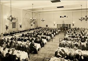 A large banquet hall with many tables and many men and women seated at each table. There are chandeliers hanging from the high ceiling. Everyone is looking up at the camera. 