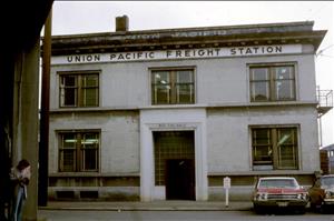 A two story concrete building with a sign along the roofline that says Union Pacific Freight Station. A red car is parked in front. A person leans against a pillar in the foreground. 