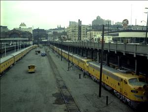Two long yellow passenger trains parked in a rail yard stretch towards the camera. The rail yard is below street level in a city. In the distance is a large brick building with the sign Union Station. Downtown buildings and billboards are visible above the rail yard.   