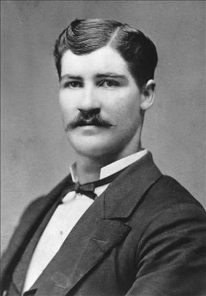 A black and white head and shoulder portrait of a white man with a handlebar mustache in a suit and cravat 