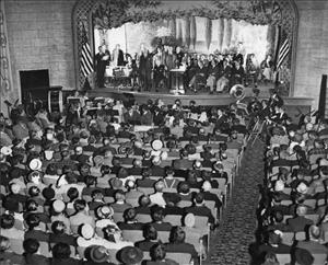 A large auditorium with all the seats filled and a youth orchestra by the stage. On the stage are a few rows of people in suits sitting and standing behind a podium. Image is black and white. 