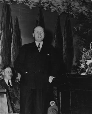 A man in a suit and tie leans against a podium smiling. Behind him is a painting of trees and another seated smiling man. The image is black and white. 