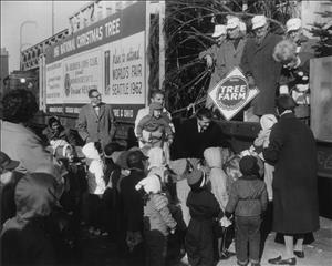 Children in winter clothing gather around a train car on which there are several adults and an evergreen tree. They are wearing white train engineer hats that say B and O on them. One of them holds a sign that says Member American Tree Farm System. A large sign on the side of the train car says 1961 National Christmas Tree