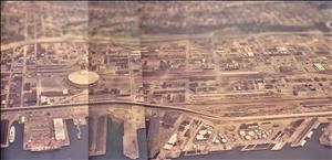 An aerial view of several cit blocks along a waterfront with piers jutting out into the water. A dome shaped stadium is on the left. A highway and train tracks meander along the water's edge. 