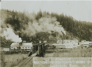 Two large buildings with smoking chimneys and various smaller ones on far side of river crossed by railroad bridge
