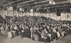 Rows and rows of people sitting in wooden chairs under a high ceiling of wooden beams. 