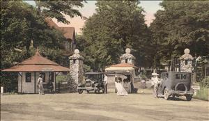 A small round building with a pagoda shaped roof next to three stone columns. Early twentieth century automobiles are passing through the columns. Men stand at the windows of the automobiles. A sign between them says STOP.  Large trees in the background. 