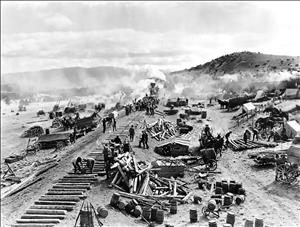 Logs are laid out next to each other in a long row at the end of which a steam locomotive waits. There are logs and tools and barrels everywhere. Workers engage in various tasks. Horses and buggies and tents dot the landscape. In the distance, rolling hills rise above the plains. 