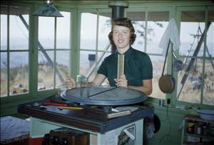 A young person stands in a room that is painted light green and has windows on all sides. They have their hands on a round, metal piece of equipment. Outside the windows are rolling hills. 