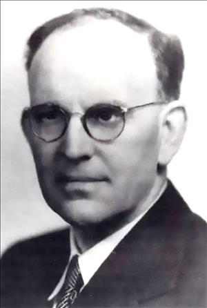 Head and shoulders portrait of a balding man in a suit and tie and round eye glasses. 