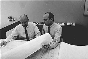Naramore and Brady sitting a a desk inside an office looking through desk sized blue prints