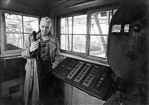 Morgan inside the control room of the bridge inspecting a mechanical device in front of a board of controls while wearing an overcoat and a plaid shirt