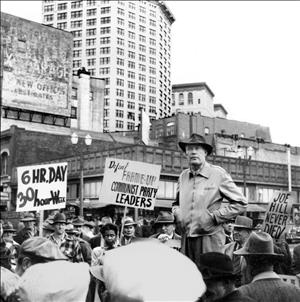 Pettus standing elevated from a crowd members holding signs that read 6 hour day, 30 hour week, Defeat Frame-up communist party leaders and Joe Hill Never Died, at a demonstration in Seattle's downtown with the Smith Tower in the background