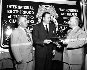 Beck receiving a wooden plaque from a man with slicked back hair and a dark suit in front of a mural that reads International Brotherhood of Teamsters... Warehousemen and Helpers of America