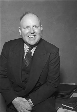 Beck smiling in a vested suit, patterned shirt and thin watch, sitting on a desk