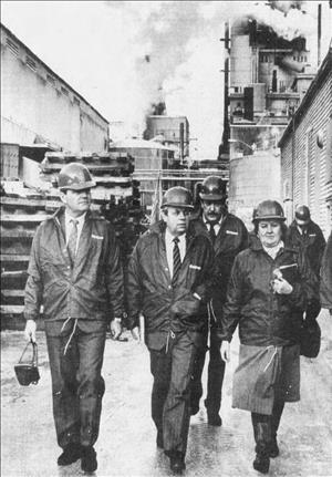 Lavroff in a slick coat over business clothes and a hardhat trailing behind three other people as they walk outside in an industrial setting
